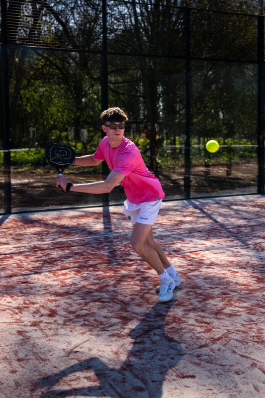 A player in a pink shirt reaching for a padel ball on a red outdoor court